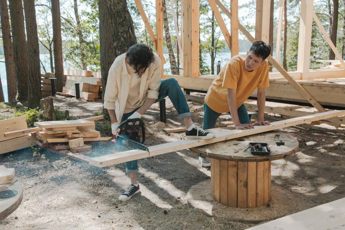 Two adult men working on wood construction project outdoors, using a chainsaw.